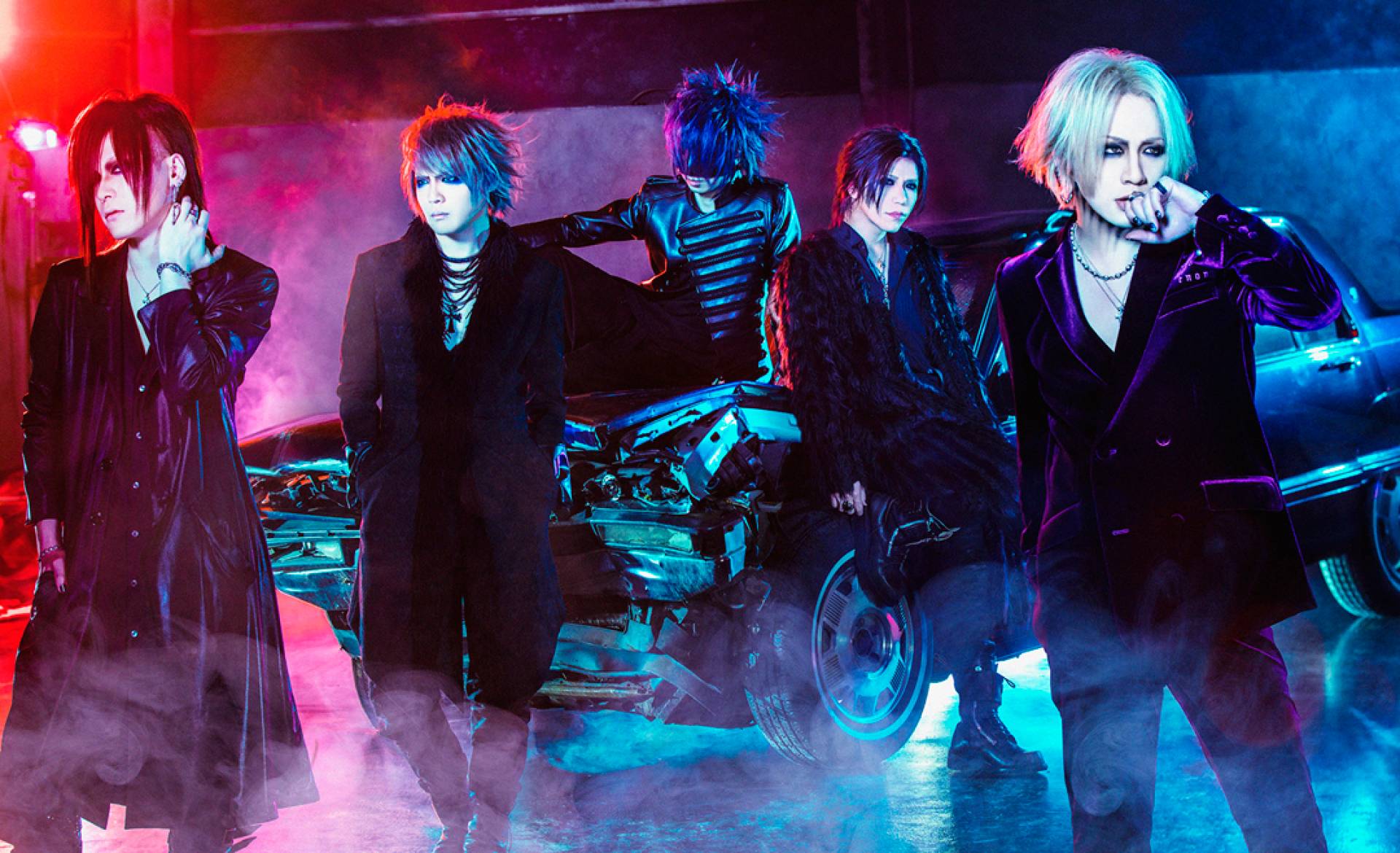 The band members of The GazettE with one of the members sitting on the bonnet of a car which has partial damage to the front of it. 